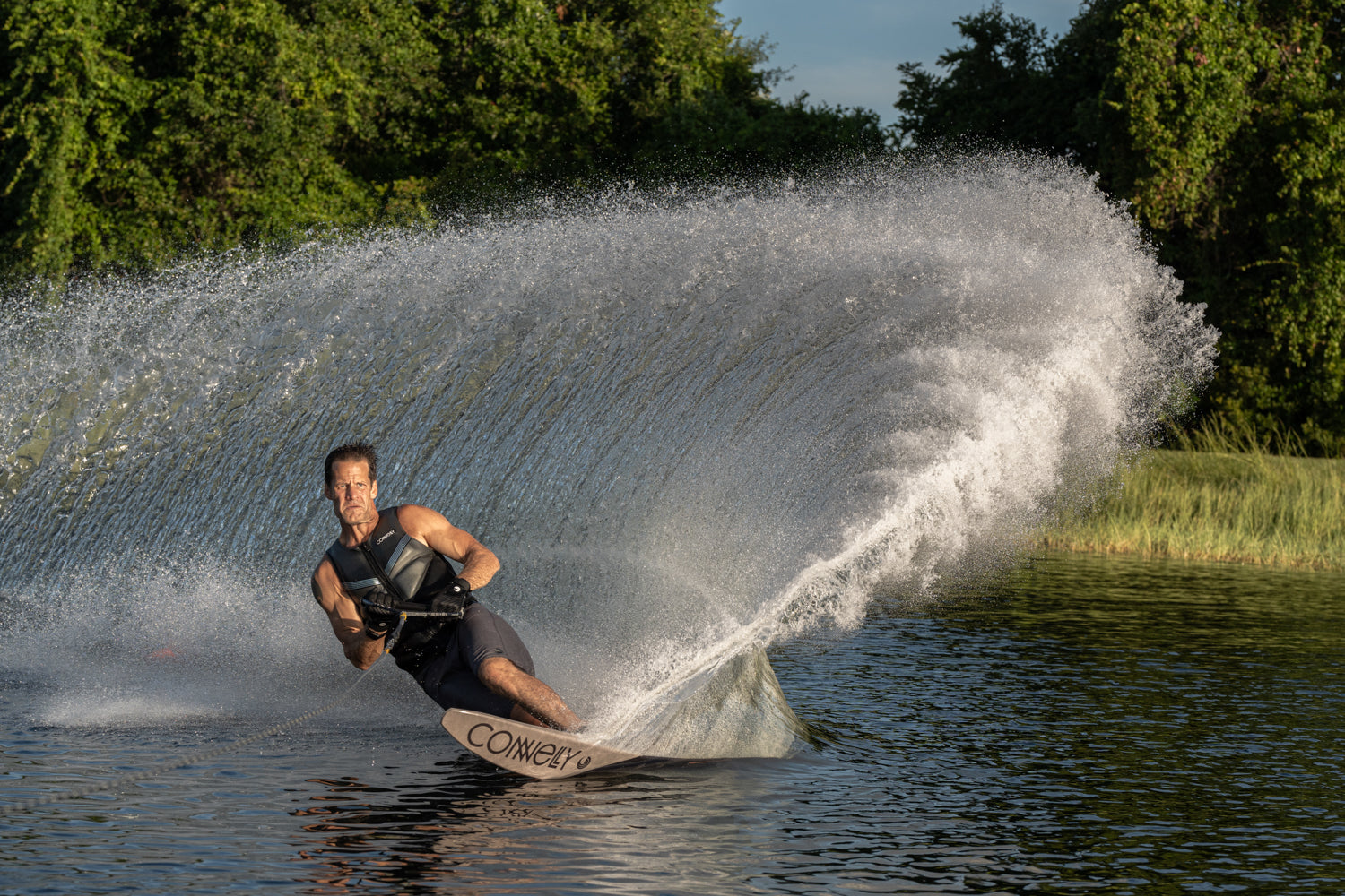Man carving a sharp turn on a Connelly water ski, creating a dramatic spray of water, with trees in the background.